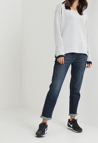 Suéter de punto gris claro con un corte holgado, acentos de puños azul oscuro; combinado con unos jeans azules oscuros arremangados y zapatillas deportivas negras.