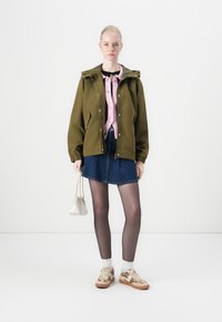 Olive green jacket with hood, pink knit cardigan, denim skirt, sheer black tights, and patterned sneakers. Complemented by a small white bag.