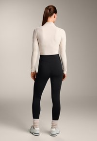 Leggings neri a vita alta con una texture liscia, abbinati a un top aderente color beige chiaro. La modella indossa calzini arricciati rosa chiaro e sneakers.