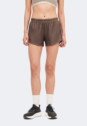 Αθλητής με καφέ σορτς Under Armour, μπεζ αθλητικό σουτιέν, λευκές κάλτσες και μαύρα παπούτσια για τρέξιμο, που στέκεται σε απλό ανοιχτό φόντο.