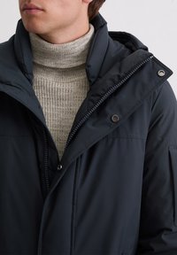 Marineblaue isolierte Jacke mit hohem Kragen, ausgestattet mit einem Reißverschluss und Druckknopfverschluss, getragen über einem hellgrauen gerippten Rollkragenpullover.