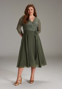 Oliven grünes Kleid mit einem Spitzenoberteil, Dreiviertelärmeln und einem fließenden Rock. Goldene Blockabsatzsandalen runden das Outfit ab.