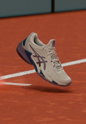 Scarpa sportiva beige chiaro e viola che solleva polvere di argilla su un campo da tennis in terra rossa vicino a una linea di confine bianca.