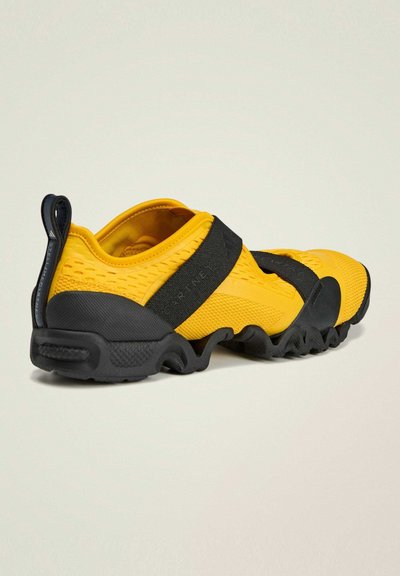 Zapatillas deportivas amarillas con parte superior texturizada, banda elástica negra y suela gruesa negra con diseño ondulado.