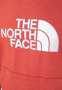 Rode sweatshirt met een groot wit logo en de tekst "THE NORTH FACE" in een gebogen ontwerp. Het materiaal lijkt zacht en textuur te hebben, met een voorkant zak.