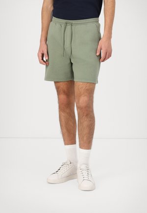 Hombre con pantalones cortos de cuerda color verde oliva, calcetines blancos, zapatillas blancas y camiseta azul marino, de pie sobre un suelo blanco frente a una pared lisa.