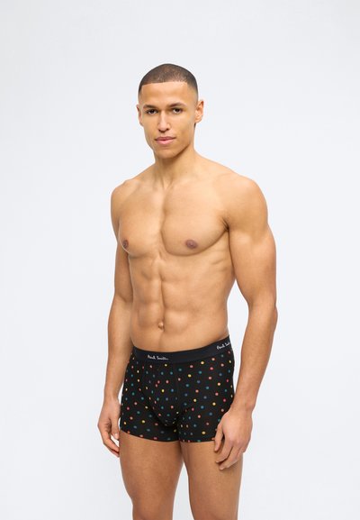 Ευδιάθετος άνδρας με κοντά μαλλιά στέκεται φορώντας μαύρα ανδρικά εσώρουχα με πολύχρωμες βούλες και ελαστική ζώνη με την ένδειξη "Paul Smith" σε απλό φόντο.