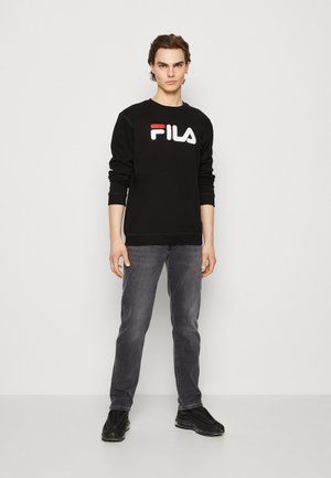 Zwart sweatshirt met Fila-logo in het wit en rood, aangegeven met donkergrijze jeans. Model staat op een effen achtergrond, met zwarte sneakers.