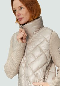 Lichtbeige gewatteerde vest met een hoge kraag, gemaakt van een gladde, glanzende stof en met ribgebreide mouwen. Ritssluiting aan de voorkant.