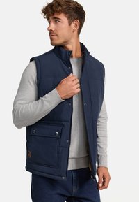 Chaleco aislante azul marino con cuello alto, botones de presión y un bolsillo frontal. Lleva puesto un suéter gris claro y jeans de mezclilla oscuros.
