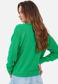 Grüner Pullover mit lockerem Schnitt, gerippten Bündchen und Saum. Weiche Textur, lange Ärmel und runder Ausschnitt. Wird über hellblauem Denim getragen.