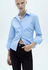 Svetlo modra srajca s kratkimi rokavi in vertikalnimi črtami, s koničastim ovratnikom in enim žepom na prsih, v kombinaciji z črnimi kavbojkami.