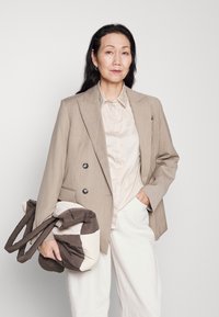 Beige dubbelknäppt kavaj med silverknappar, tillsammans med en ljus satinblus och vida vita byxor. Håller en vadderad brun och krämfärgad väska.