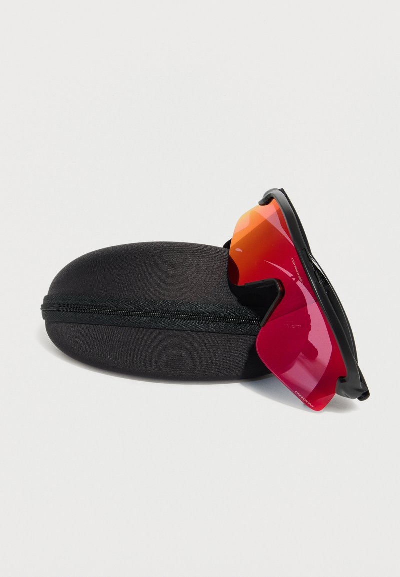 Lunettes de sport avec des verres rouge-orange reposant sur un étui protecteur noir à fermeture éclair sur fond blanc.