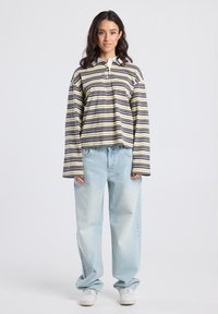 Nuori nainen seisoo, päällään pitkähihainen raidallinen t-paita, vaaleansiniset löysät farkut ja valkoiset lenkkarit, neutraali tausta.