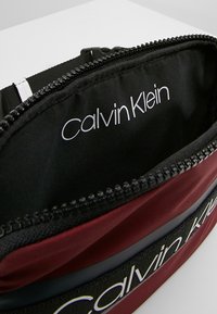 Calvin Klein heuptasje met een kastanjebruine buitenkant en zwarte voering, voorzien van een opvallend logo aan de binnenkant en een ritssluiting.