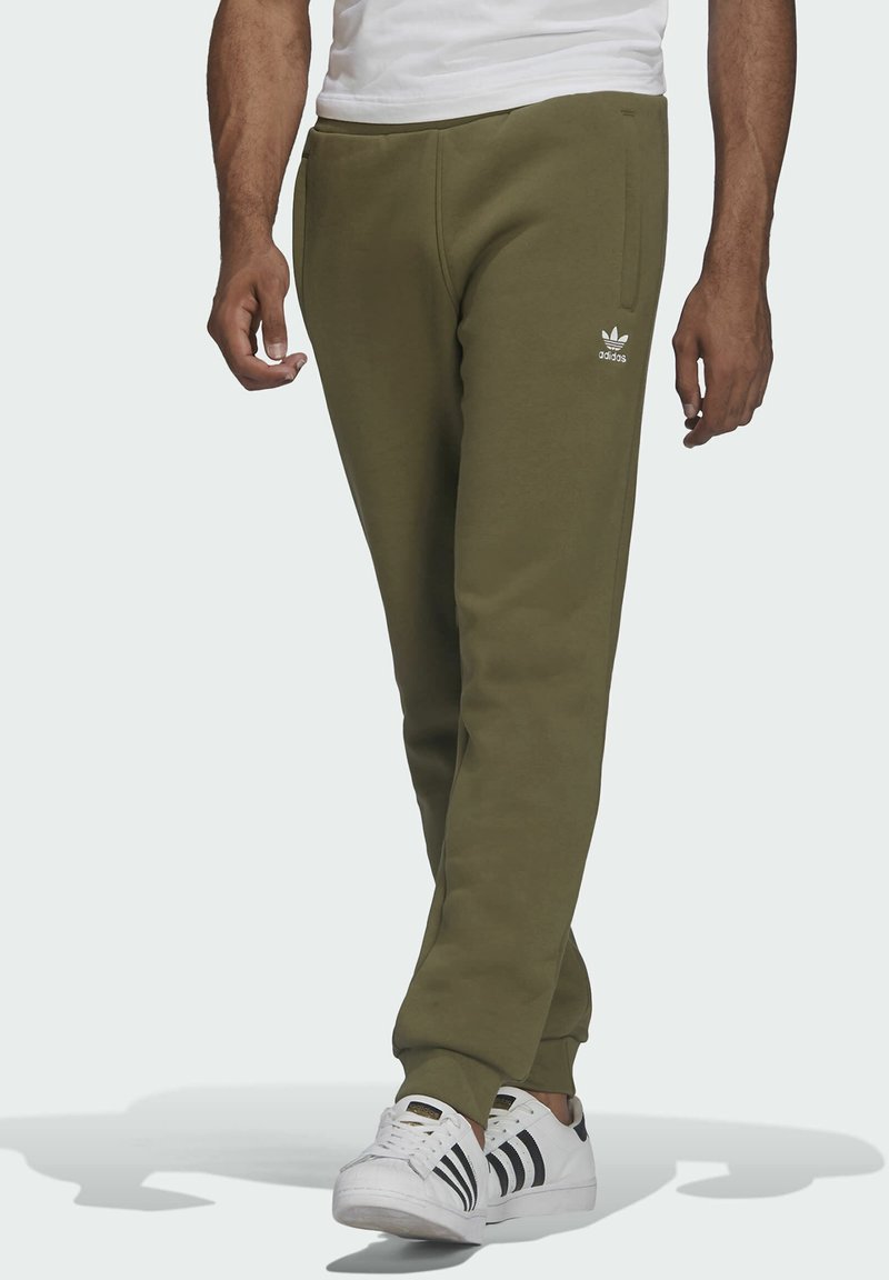 Olivgröna mjukisbyxor med elastiska muddar, sidofickor och en vit Adidas-logotyp på vänster lår. Kombinerade med vita sneakers med svarta ränder.