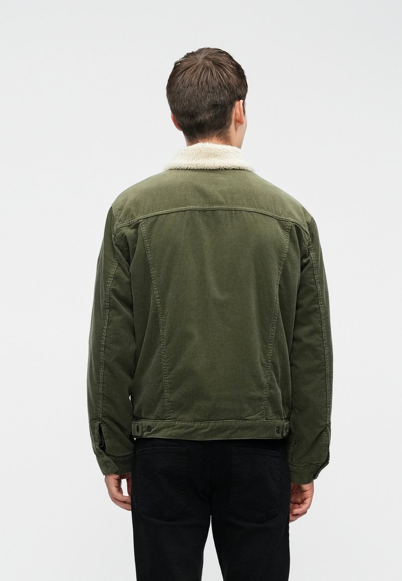 Homme de dos portant une veste en velours côtelé vert olive avec un col sherpa blanc et un pantalon noir sur fond uni.