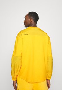 Gul langærmet skjorte med en afslappet pasform, der har kontrasterende stofærmer og et logo på ryggen. Glat tekstur, enkel design.