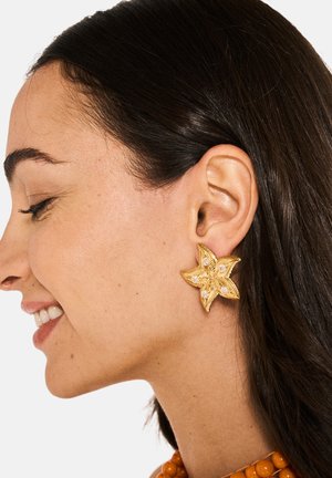 Boucles d'oreilles florales en or avec un design texturé et des gemmes claires incrustées, mettant en valeur de délicates pétales en forme de feuille sur un teint de peau.