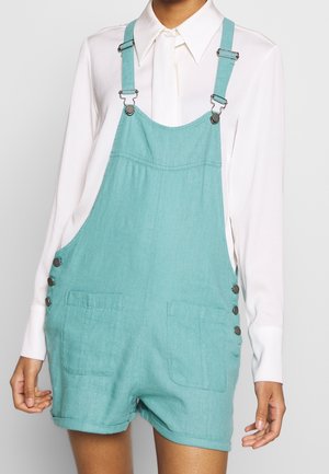 Personne portant une salopette courte en denim bleu sarcelle avec des détails à boutons sur les côtés, par-dessus une chemise blanche à manches longues et col, debout devant un fond uni.