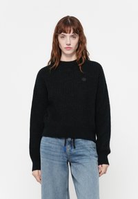 Zwarte gebreide trui met een ronde hals en verlaagde schouders. Heeft een logo patch en een trekkoord aan de onderkant, gecombineerd met blauwe jeans.