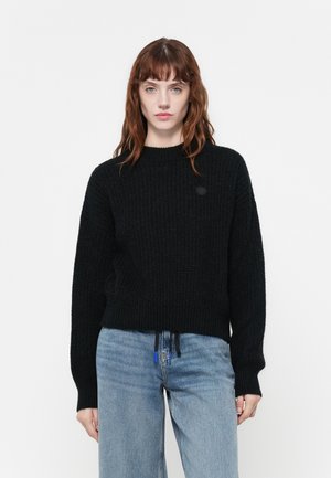 Black knitted sweater with a round neckline and drop shoulders. Features a logo patch and drawstring at the hem, paired with blue jeans.