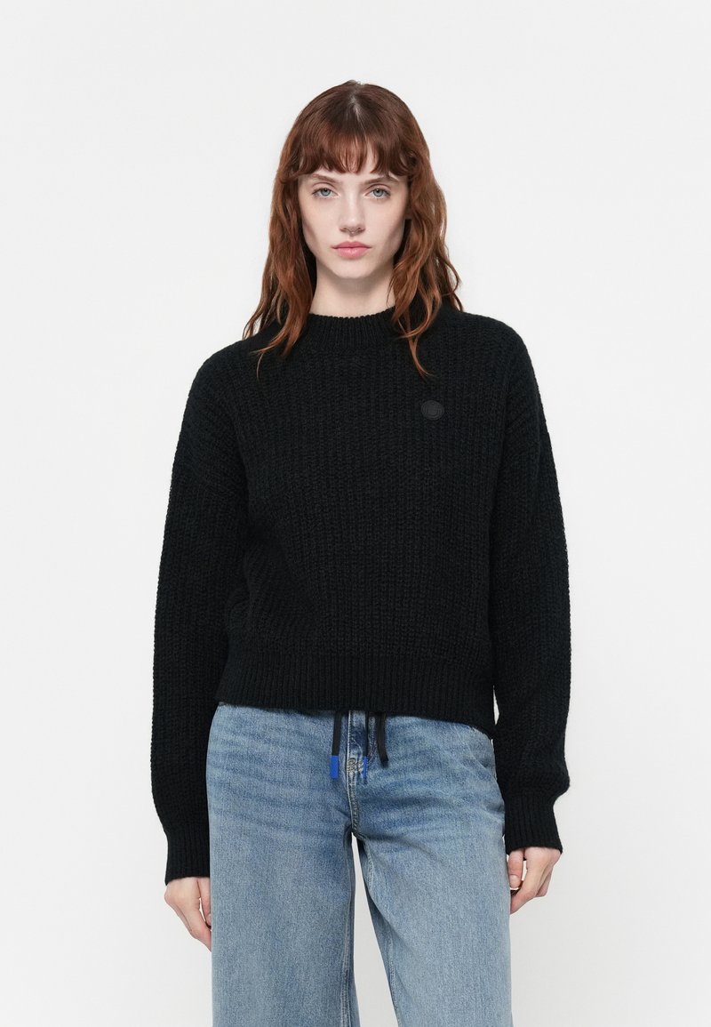 Schwarzer Strickpullover mit rundem Ausschnitt und überschnittenen Schultern. Mit einem Logo-Patch und Kordelzug am Saum, kombiniert mit blauen Jeans.