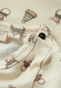 Un tissu crème côtelé présente de petits motifs d'ours bruns et de glands. L'encolure possède une fermeture à bouton et une texture douce et lisse.
