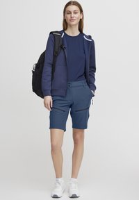 Sweat à zip navy sur un t-shirt navy, associé à un short bleu avec deux poches zippées et une texture lisse. Fonctionnalité mise en avant.