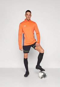 Mees oranžis sporditellis, mustades lühikarides ja sokkides seisab üks jalg jalgpallil, lihtsa valge tausta ees.