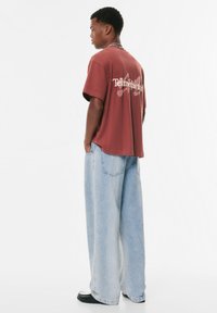 T-shirt graphique bordeaux avec le texte "Tell Me", associé à un jean en denim bleu clair à jambes larges. Modèle positionné de profil, mains dans les poches.