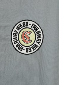Šedá tkanina s vyšívanou kulatou nášivkou. Nášivka obsahuje žlutý a červený design s textem "ODCHÁZÍME - DALÉKO" v černé barvě.