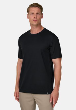 Homme portant un t-shirt noir uni à manches courtes et un pantalon beige, debout devant un fond clair uni.