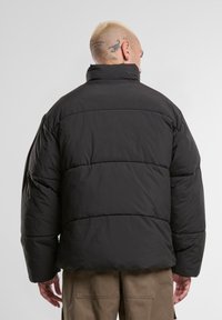 Homme aux cheveux blonds courts et tatouages sur la tête, portant une veste en doudoune noire et un pantalon marron, tourné vers le dos devant un fond uni.