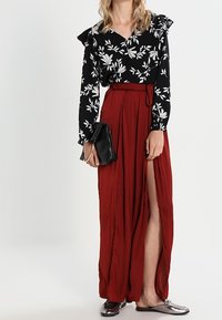 Blusa nera floreale con maniche a sbuffo, abbinata a una lunga gonna rossa fluida con uno spacco alto. Infradito argentati e pochette nera.