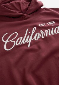 Bordeauxrode hoodie van zachte stof met geborduurde witte tekst "California" en hierboven in eenvoudige letters "EST 1989" gedrukt.