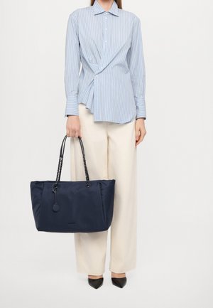 Femme portant une blouse rayée bleu clair et un pantalon crème, tenant un grand sac fourre-tout bleu marine avec des poignées en chaîne, se tenant devant un fond uni.