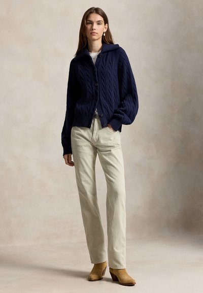 Femme portant un cardigan bleu marine en maille torsadée boutonné, un pantalon beige et des bottines marron clair, debout devant un fond beige uni.