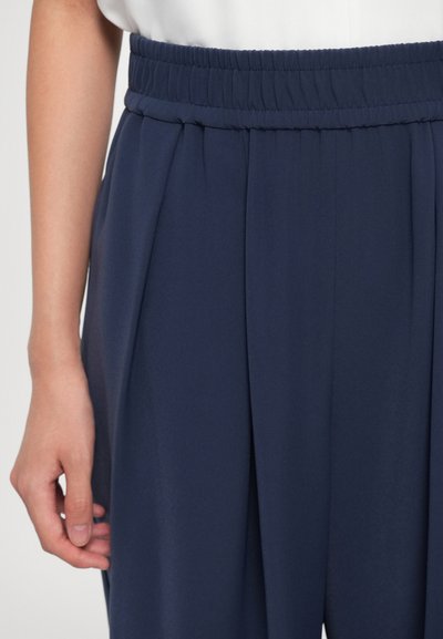 Left hand relaxed beside navy blue high-waisted pants with gathered elastic waistband and front pleats, on a person wearing a white top.