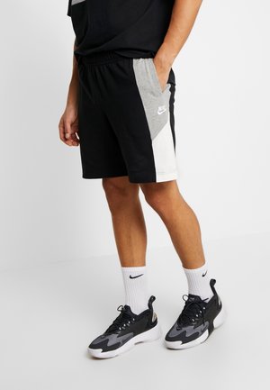 Man draagt zwarte, grijze en witte Nike-short met witte Nike-sokken en zwarte, grijze en witte Nike-sneakers, staand tegen een witte achtergrond.