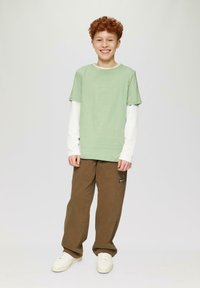 Glunderende jongen met rood haar, gekleed in gelaagde groene en witte langesleeveshirts, bruine broek en witte sneakers, staand tegen een effen achtergrond.
