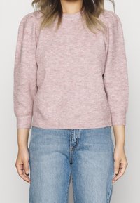 Jasnoróżowy prążkowany sweter z okrągłym dekoltem i trój-ćwiartkowymi bufkami, zestawiony z niebieskimi dżinsami. Miękka tekstura i luźny krój.