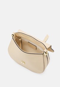 Beżowa skórzana torba na ramię z klapą, zapięciem w odcieniu złota, wewnętrzną kieszenią na zamek oraz małą pętlą na pasek na jednolitym białym tle.