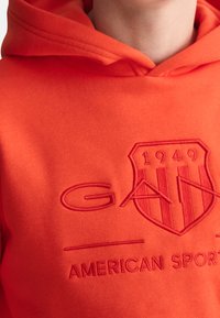 Orange Kapuzenpullover aus weichem Stoff mit einem auffälligen geprägten Wappen-Design, das die Schriftzüge "GAN", "1949" und "AMERICAN SPORTS" enthält.