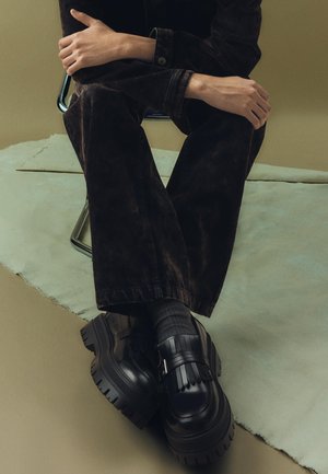 Tömbös fekete platform cipők rojtos pánttal, sötét, texturált, bő szárú nadrággal és szürke zoknival párosítva, semleges háttér előtt.