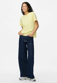 Gul kortärmad T-shirt med vita prickar, kombinerad med vida mörkblå denimjeans. Modellen står med händerna i fickorna.