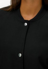 Gros plan sur une personne portant une veste noire à boutons avec deux boutons-pression en argent visibles et un col côtelé.