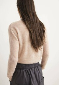 Hellrosa strukturierter Strickpullover mit langen Ärmeln; kombiniert mit dunkler Hose mit elastischem Bund. Das Haar fällt bis zur Mitte des Rückens und verschmilzt mit dem neutralen Hintergrund.