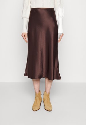 Femme portant une jupe midi marron brillante, un haut blanc à manches longues et des bottines en daim beige, debout sur un sol clair et un fond uni.